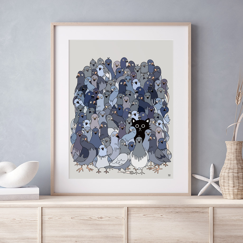 Framed illustrated art print of a large flock of grey and blue pigeons with a single black cat hidden among them, displayed in a light wood frame on a pale wooden sideboard with neutral home décor accessories.