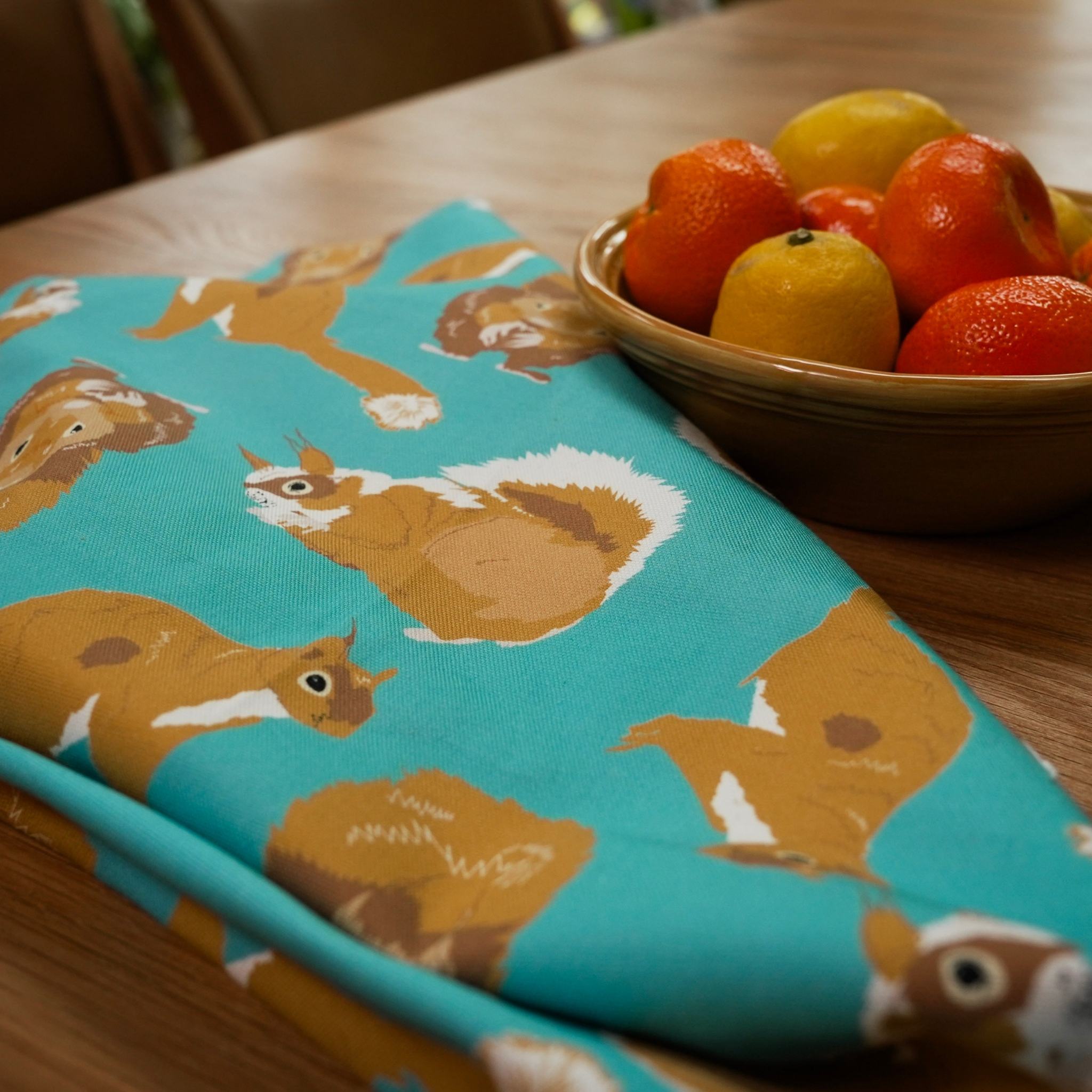 Red Squirrel design teal cotton tea towel shown on table next to fruit bowl