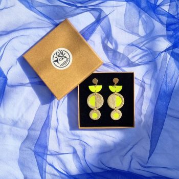Geometric acid yellow and gold leather dangly earrings featuring a circular inlay design, in a kraft gift box resting on a draped bright blue tulle fabric background.