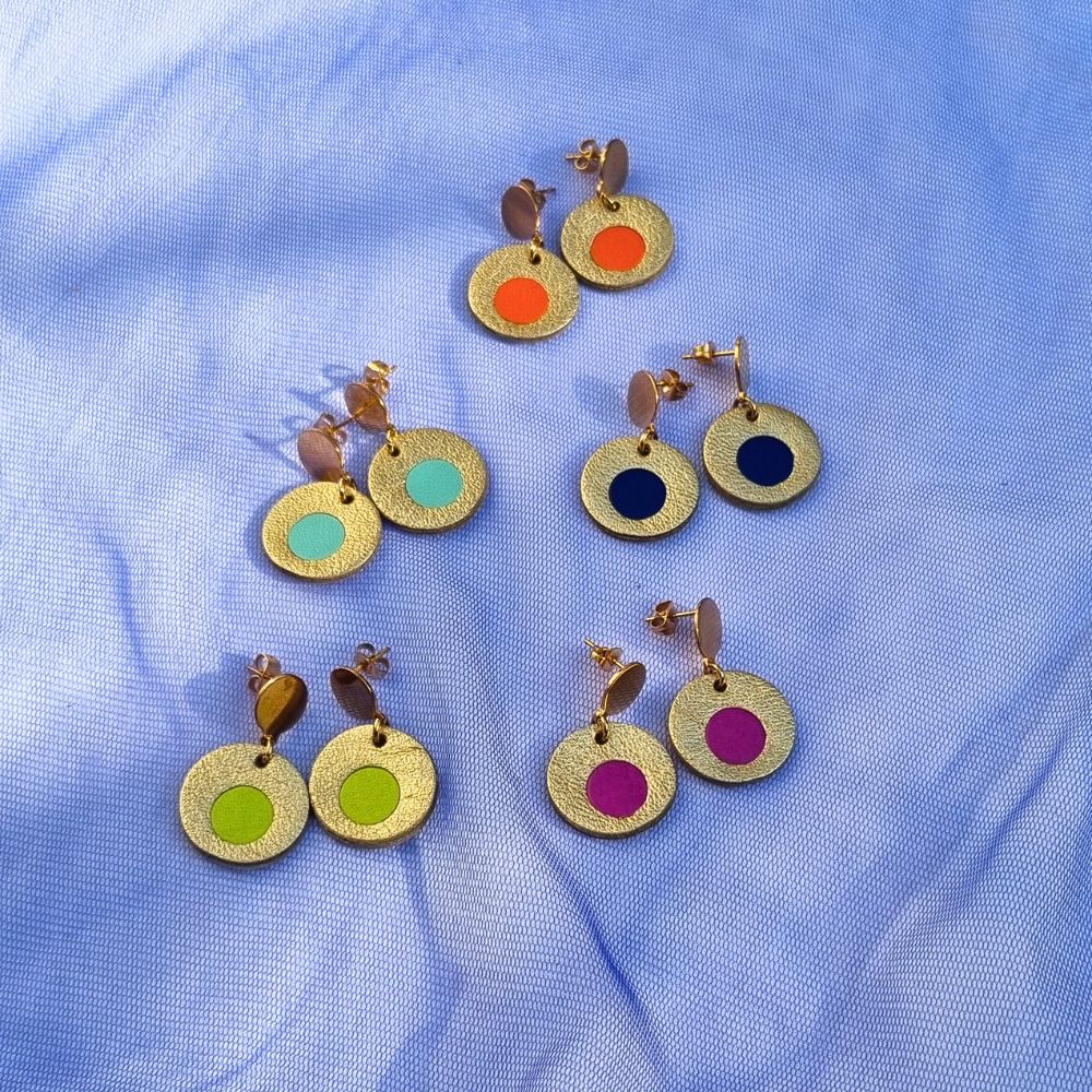Five pairs of circular leather drop earrings in gold, featuring colourful centre inlays in orange, mint, navy, lime, and magenta, arranged on blue mesh fabric.